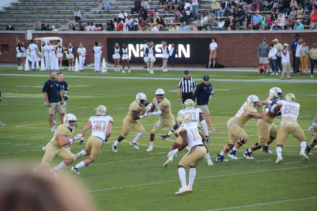 Menschen in Sportkleidung und Helmen spielen Fußball auf einem Feld, während andere Kameras halten, mit Zuschauern auf Bänken und Text auf einer Hintergrundwand.