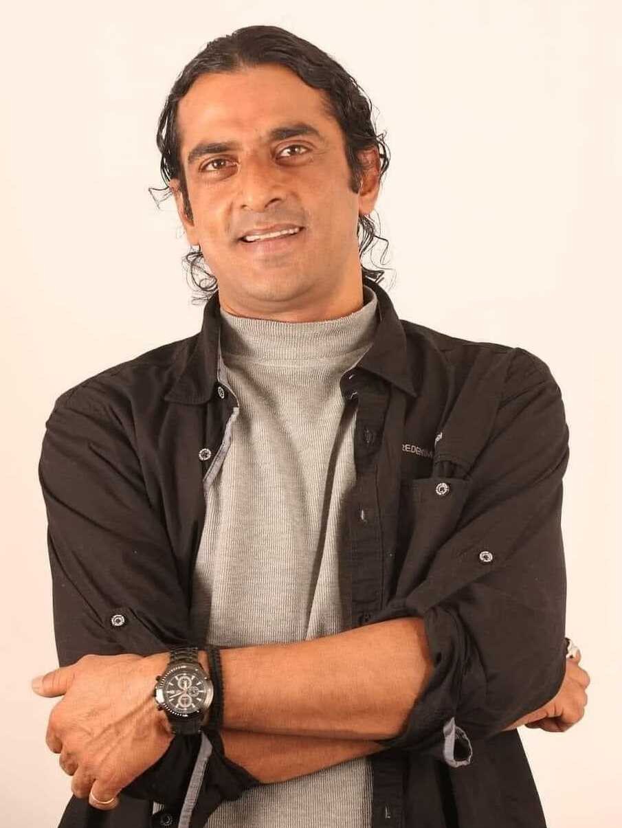 Ein Mann in einem schwarzen und grauen Anzug mit überschlagenen Armen, der eine Uhr tr├Ągt und gegen einen weißen Hintergrund gestellt ist, der nachdenklich erscheint.