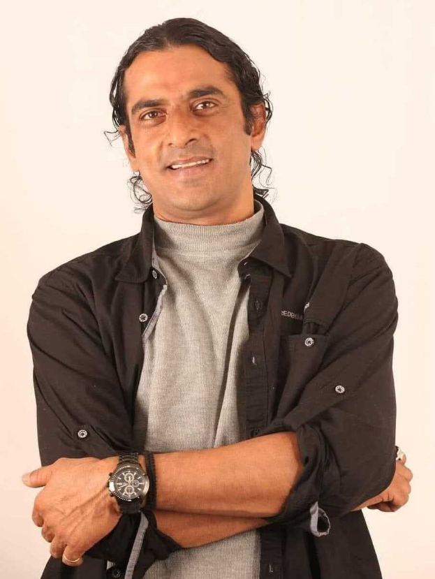 Ein Mann in einem schwarzen und grauen Anzug mit überschlagenen Armen, der eine Uhr tr├Ągt und gegen einen weißen Hintergrund gestellt ist, der nachdenklich erscheint.