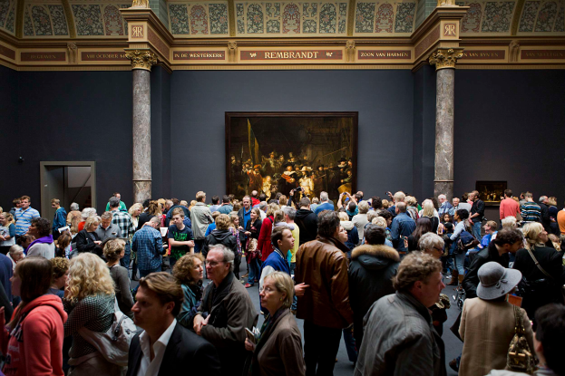 Eine große Gruppe von Menschen in einem Raum mit einem Foto auf der Wand, Säulen, einer Tür und einer Decke mit Lampen, einige halten Taschen, im Stedelijk Museum Amsterdam.