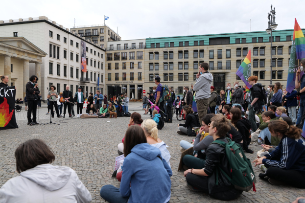 Eine Gruppe von Menschen, die auf dem Boden vor einer Menge sitzen, die Fahnen und Transparente hält, mit einer Person, die in ein Mikrofon spricht, einer Statue und Gebäuden im Hintergrund während einer anti-schwulen Demonstration in Berlin.
