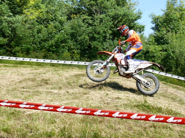 Eine Person fährt ein Fahrrad und trägt einen Helm, umgeben von Absperrband, mit Bäumen, Gras und Himmel im Hintergrund.