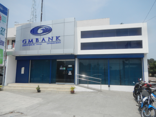 Ein modernes Gebäude mit Glaswänden und -türen, identifiziert als Gmbank Bank in Hyderabad, mit Treppen, Geländern, Topfpflanzen, Fahrzeugen auf der Straße, einem Banner mit Text, einem Turm mit Drähten, Bäumen und einem bewölkten Himmel im Hintergrund.