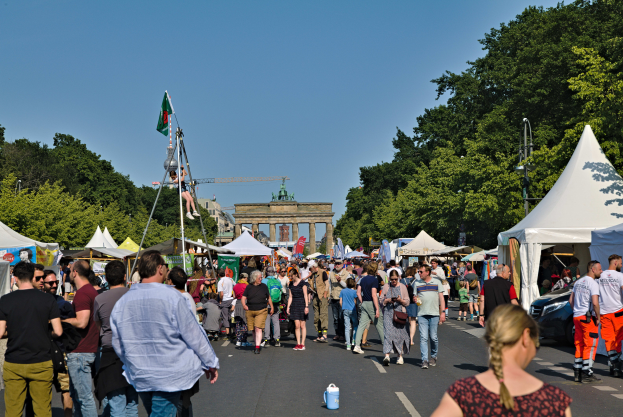Eine Menschenmenge, die eine Straße mit Zelten, Fahrzeugen und Bäumen entlanggeht, mit einem Bogen und einem klaren blauen Himmel im Hintergrund und Polen mit Flaggen auf der linken Seite, die wahrscheinlich das Oktoberfest in München, Deutschland, darstellen.