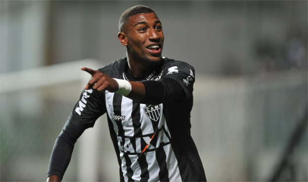 Ein Fußballspieler in einem schwarz-weißen gestreiften Trikot feiert mit einem Lächeln nach einem Torerfolg, der Hintergrund unscharf, um den Moment zu betonen.