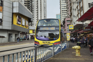 Ein gelber Doppeldeckerbus fährt auf einer Straße mit hohen Gebäuden entlang, mit Fußgängern auf der rechten Seite, einem Hydranten und einem Geländer, unter einem klaren blauen Himmel.
