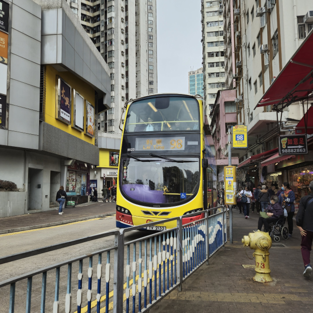 Ein gelber Doppeldeckerbus fährt auf einer Straße mit hohen Gebäuden entlang, mit Fußgängern auf der rechten Seite, einem Hydranten und einem Geländer, unter einem klaren blauen Himmel.