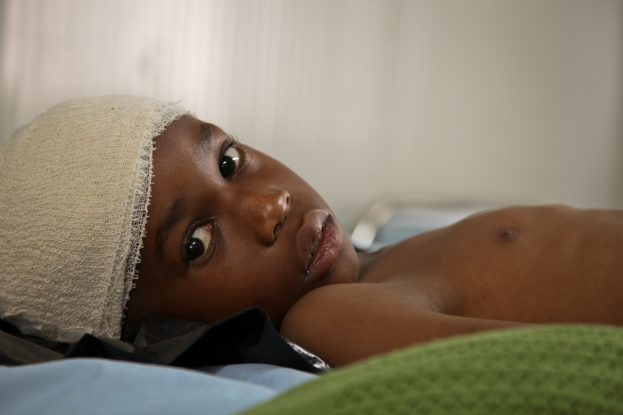 Ein junger Junge liegt in einem Krankenhausbett mit einem Verband auf dem Kopf, bedeckt von einer Decke und einem grünen Tuch, mit einem unscharfen Hintergrund.