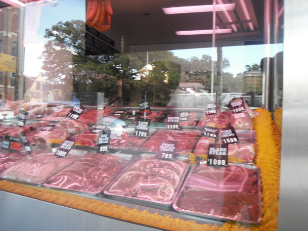 Eine Glasvitrine in einem Fleischgeschäft, die Tabletts mit Fleisch und Preisschildern zeigt, mit Gebäuden und Bäumen durch das Fenster und der Decke des Raums oben im Bild.