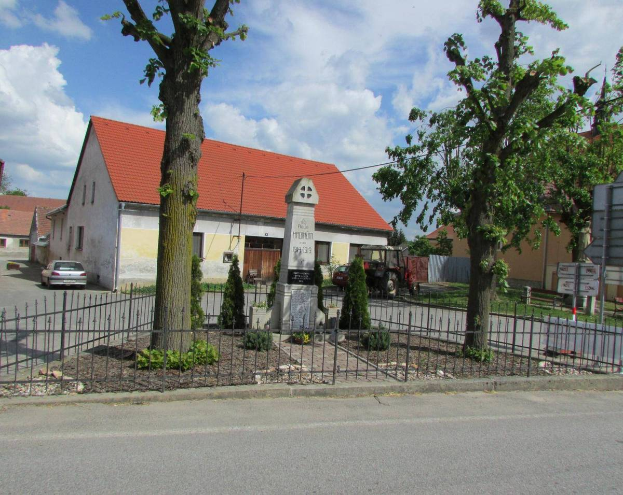Eine Kleinstadt-Szene mit der Kirche des Heiligen Grabes in St. Vitus im Zentrum, umgeben von einem Metallzaun, Bäumen, Pflanzen, Gras, einem Schild, Fahrzeugen, Polen, Drähten und einem bewölkten Himmel.