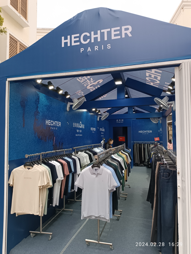 Ein Hechter Paris Stand auf der Paris Fashion Week, mit präsentierter Kleidung, einer Person, Lichtern, einem Gebäude und einem Baum, mit sichtbarem Himmel und einem Wasserzeichen in der rechten unteren Ecke.