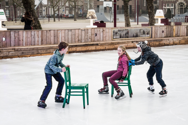 Kinder beim Skifahren im Vordergrund eines Spielplatzes mit drei Kindern und zwei Stühlen in der Mitte und Gebäuden, Bäumen, Bänken, Pfosten und einem Basketballfeld im Hintergrund.