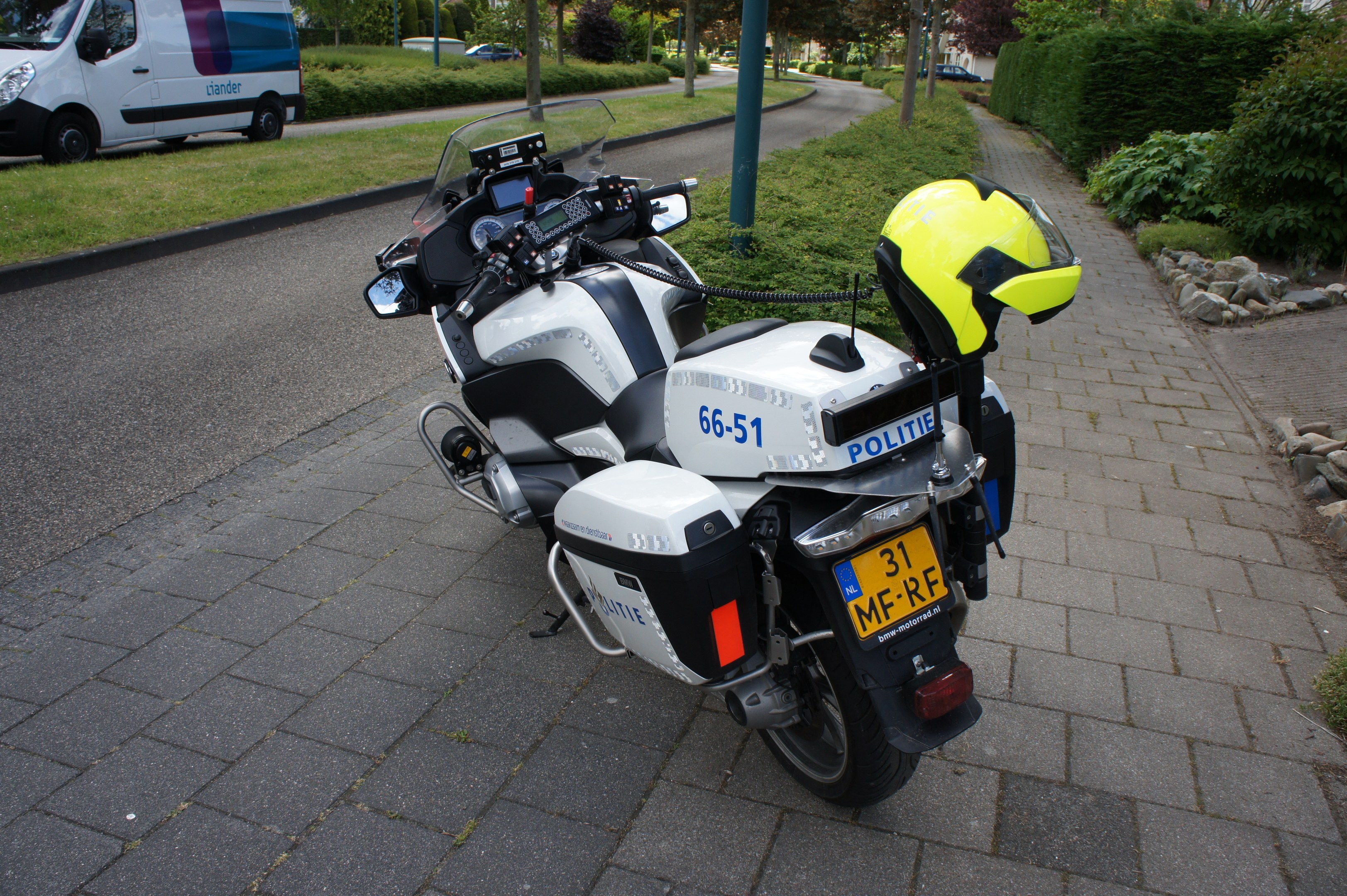 Ein Polizeimotorrad am Straßenrand mit einem Helm darauf, umgeben von Bäumen, Pfählen, Gebäuden, Pflanzen, Gras, Steinen und anderen Fahrzeugen im Hintergrund.