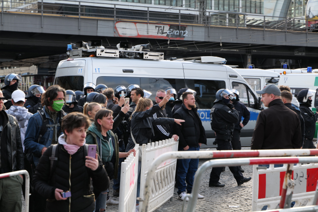 Eine Gruppe von Menschen steht hinter Barrikaden und schaut auf eine Reihe von Polizeiwagen, einige tragen Helme und halten Telefone, mit einer Brücke und Gebäuden im Hintergrund während einer Demonstration in Berlin.