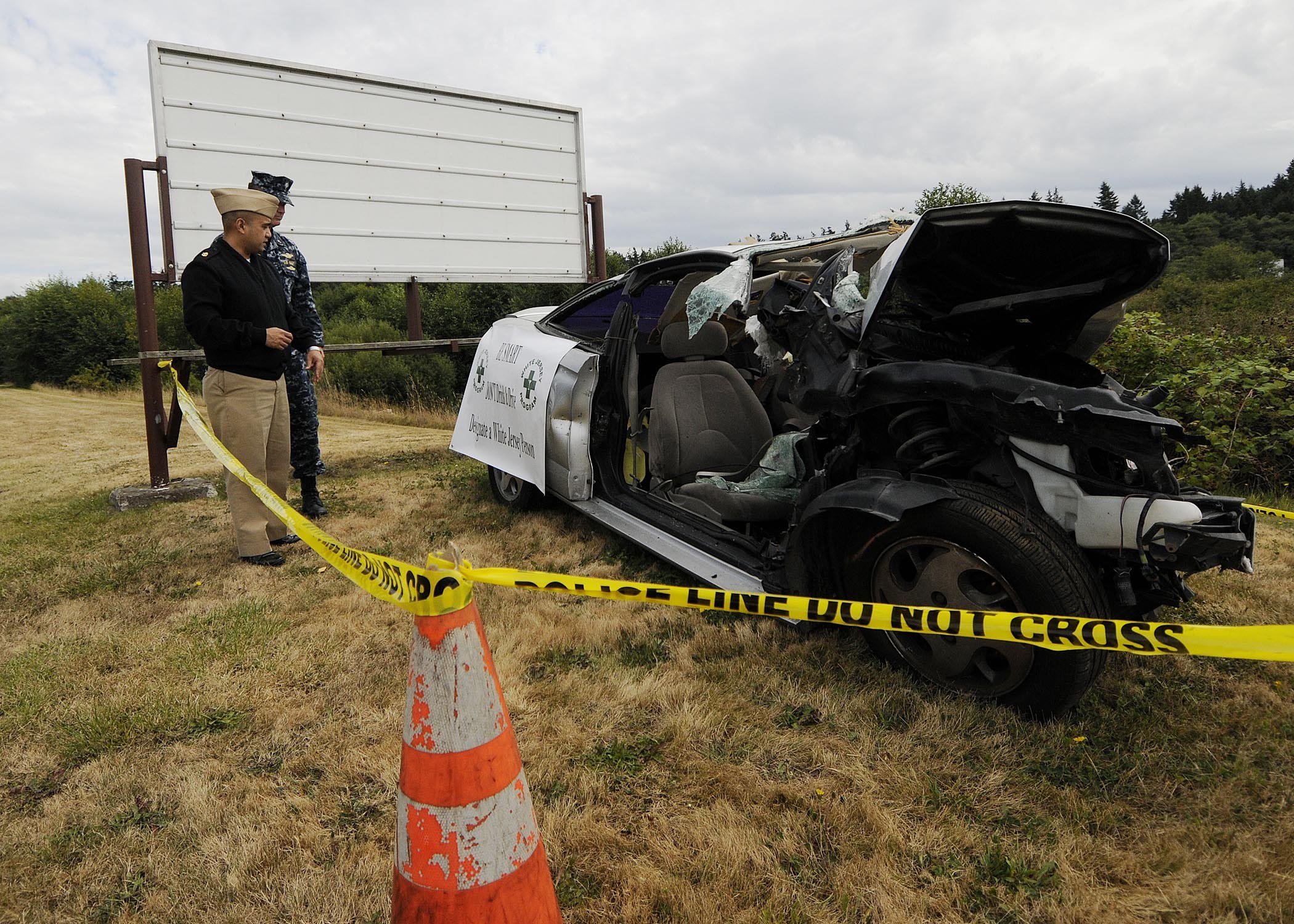 Polizeibeamter in Uniform neben einem schwer beschädigten Auto am Straßenrand mit eingedrückter Haube und zersplitterten Fenstern, neben einem Verkehrskegel und einem bandagierten Pfahl, mit Bäumen und einem bewölktem Himmel im Hintergrund.