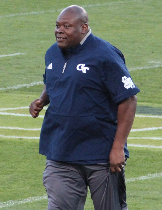 Ein Mann in einem blauen Hemd und grauen Hosen steht auf einem grünen Feld, lächelt mit einem selbstbewussten und entschlossenen Gesichtsausdruck, was darauf hindeutet, dass er ein Fußballtrainer ist.