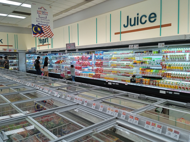 Ein hell erleuchteter Supermarktgang mit Flaschengetränken, frischem Obst und anderen Lebensmitteln, mit einigen Menschen, die durch die Gänge schlendern und einer deckenmontierten beleuchteten Tafel.