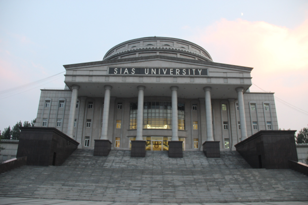 Großes Gebäude mit Säulen, Fenstern, Türen und Treppen, umgeben von Bäumen unter einem klaren blauen Himmel, mit einem Universitätsnamen auf der Fassade.
