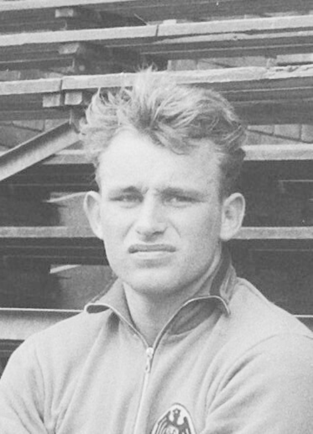 Ein Schwarz-Weiß- Foto eines Mannes in einer Jacke, der vor einem Haufen Holzplanken steht, der für den legendären deutschen Fussballer gehalten wird.