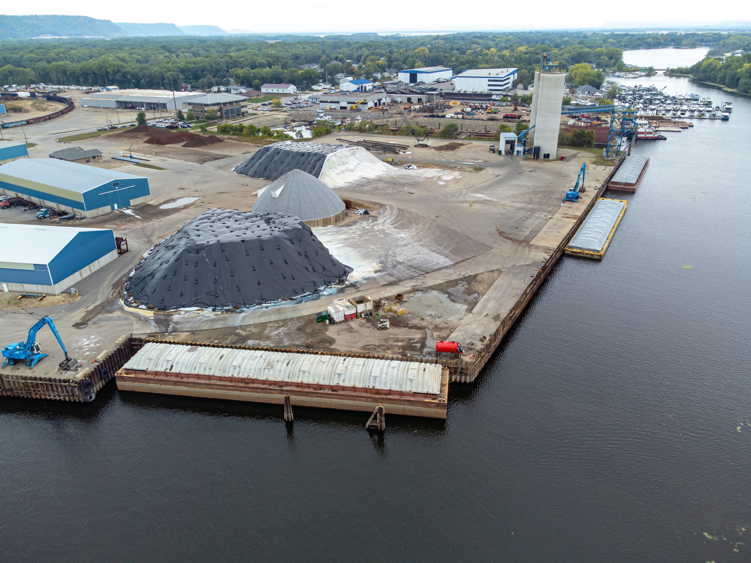 Luftaufnahme eines großen Gewässers mit einem zentralen Dock, umgeben von Gebäuden, Schuppen, Fahrzeugen, Kränen und anderen Strukturen, mit Bäumen, Hügeln und einem klaren blauen Himmel im Hintergrund.