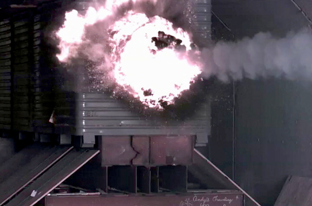 Eine große Explosion in der Mitte eines Gebäudes mit Rauch, einer Wand im Hintergrund und Metallobjekten mit einer Tafel unten.