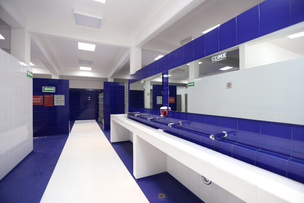 Öffentliche Toilette mit blauen Kacheln an den Wänden, weißen Countertops, Waschbecken mit Hähnen, einer Plattform mit einer Flasche und anderen Gegenständen, Informationsschildern an den Wänden, Deckenleuchten und tragenden Säulen.