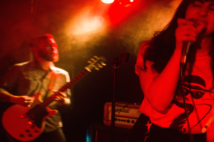 Eine Frau singt in ein Mikrofon auf der Bühne, während ein Mann im Hintergrund Gitarre spielt und Deckenlampen die lebendige Szenerie beleuchten.