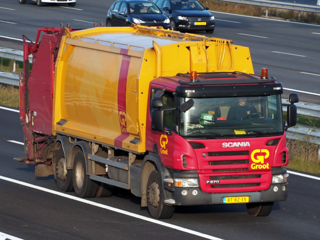 Ein gelber und roter Mülllaster fährt auf einer Autobahn, umgeben von Autos, mit einer Begränzung und Gras auf beiden Seiten der Straße.