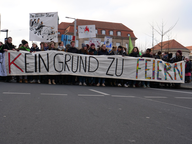 Demonstranten mit einem Banner "Kein Grund zu Feiern" gegen deutsche Sparmaßnahmen, mit Straßeninfrastruktur und Gebäuden im Hintergrund bei klarem Himmel.