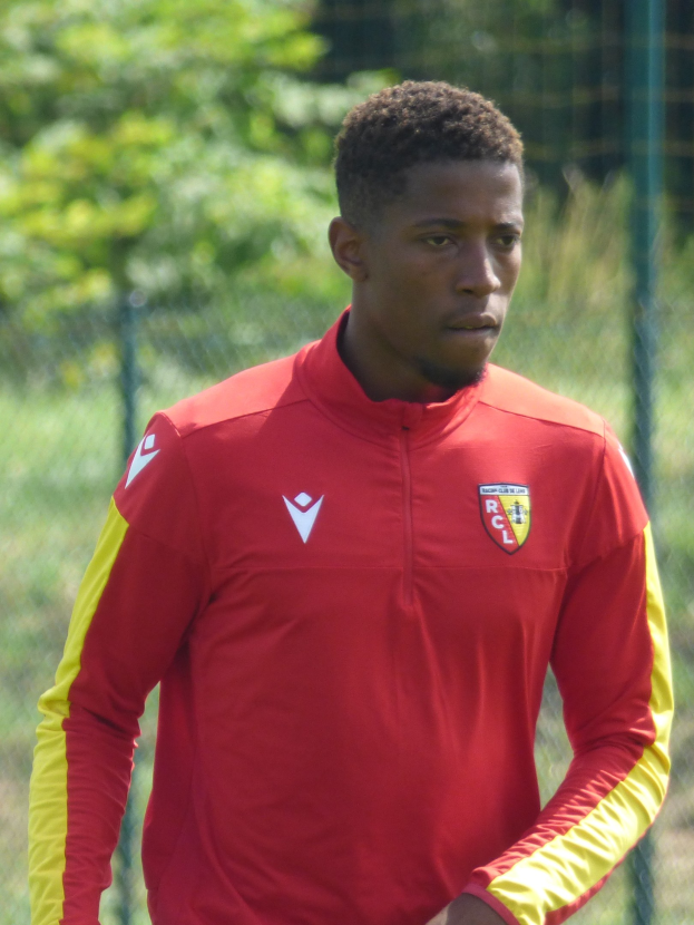 Ein junger Mann in einem roten und gelben Fußballtrikot steht auf einem Feld, hält einen Ball und hat einen Zaun und Bäume im Hintergrund.