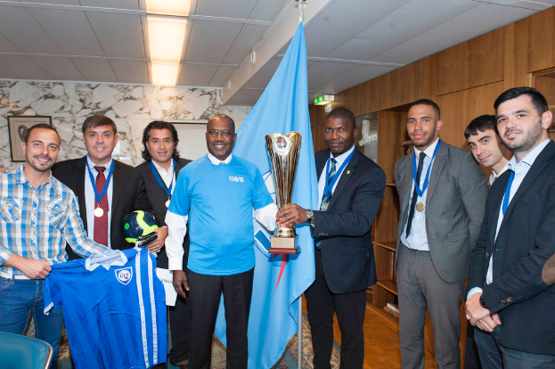 Eine Gruppe von Menschen, die zusammenstehen, wobei zwei in der Mitte einen Pokal halten, einer links ein T-Shirt und ein anderer links einen Ball hält, sowie eine Flagge im Hintergrund.