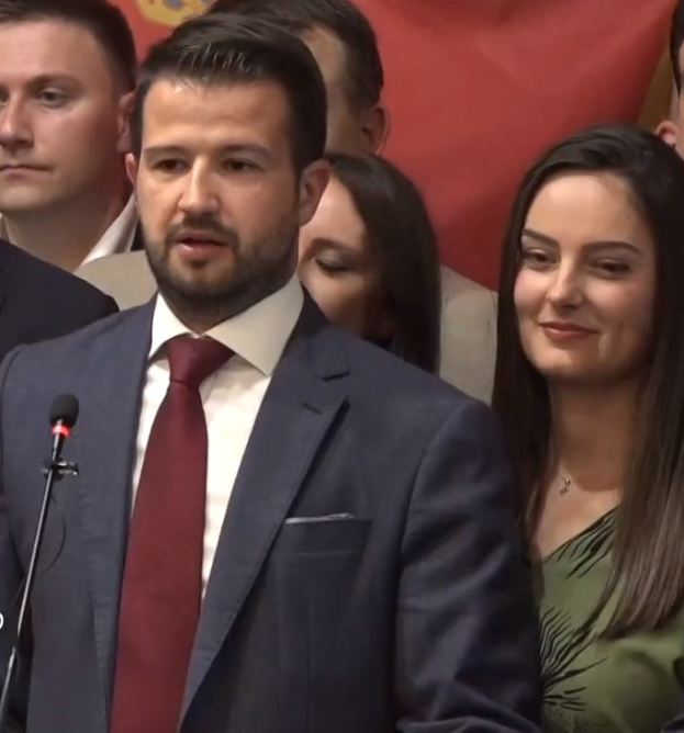 Ein Mann im Anzug spricht in ein Mikrofon an einem Podium, mit einem Banner dahinter und einer Menge davor, wahrscheinlich zu einem Regierungsverbot von Social Media.