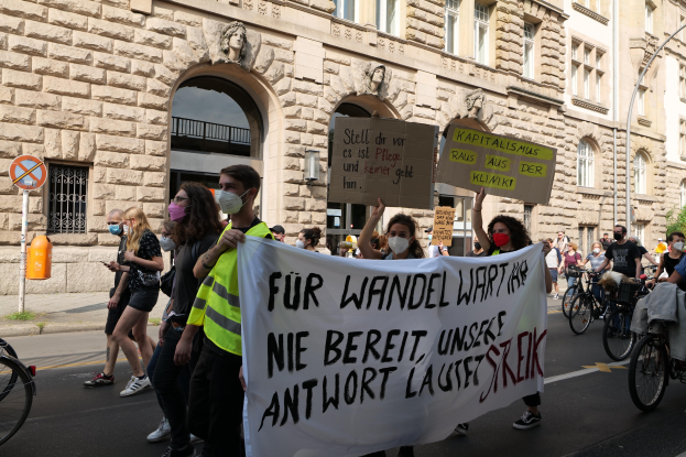 Eine Gruppe von Menschen marschiert auf einer Stadtstraße bei einer Protestaktion, einige halten Schilder und andere fahren Fahrräder, im Hintergrund ein historisches Gebäude und Bäume.