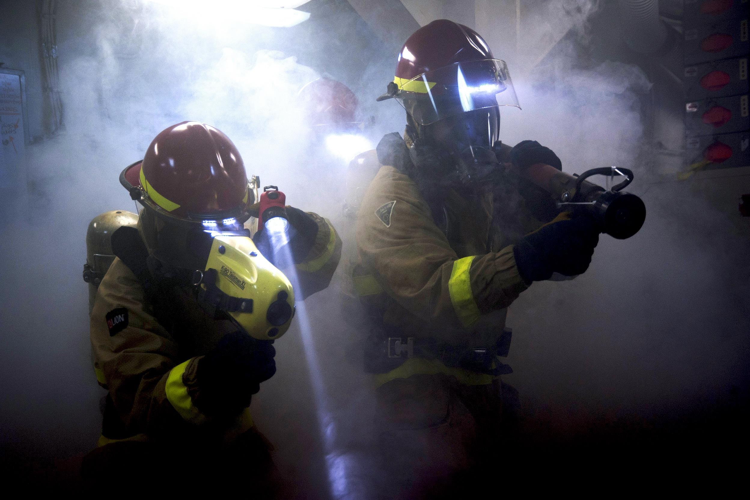 Zwei Feuerwehrleute in Schutzausrüstung sprühen Wasser auf eine rauchgefüllte Wand, mit einer Tafel und verschiedenen Objekten in der Nähe.