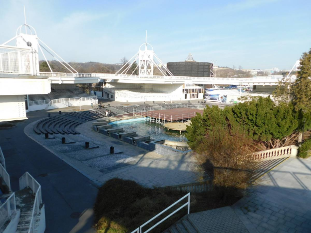 Ein Blick auf das Olympische Stadion von einem hohen Standpunkt aus, mit verschiedenen Objekten und Grün im Vordergrund und einem bewölkten Himmel im Hintergrund.