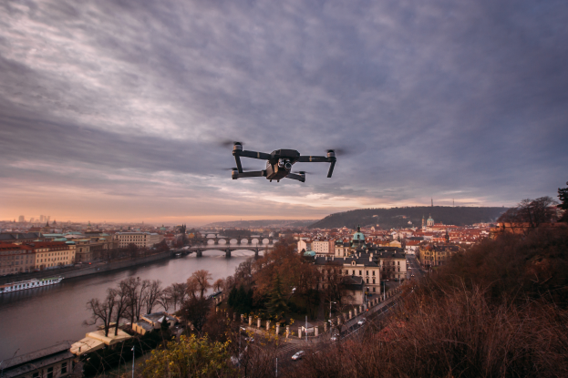 Ein Drohne, das über einer Stadt mit Gebäuden, Bäumen und Fahrzeugen fliegt, mit einem Fluss und einer Brücke im Hintergrund.