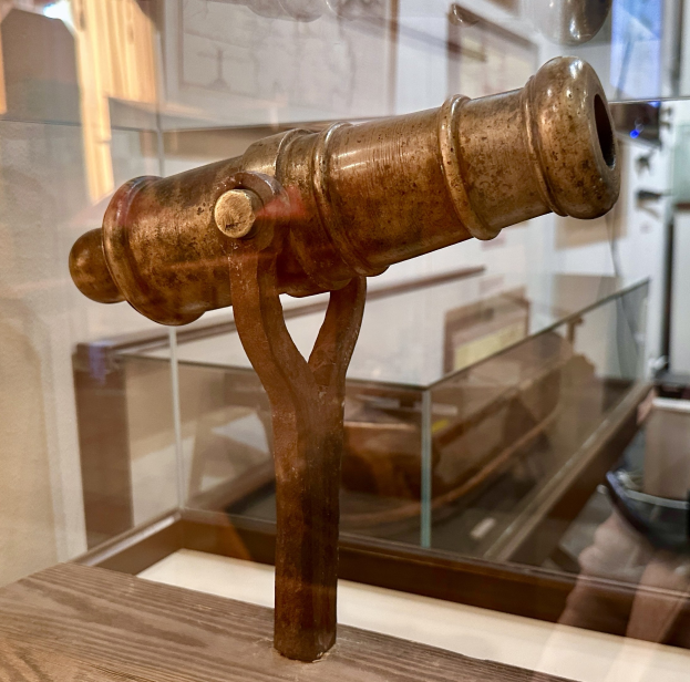 Alte Messingkanone in einer Glasvitrine mit einem Holzunterbau, vor einer Wand mit Rahmen in einem Museums Setting.