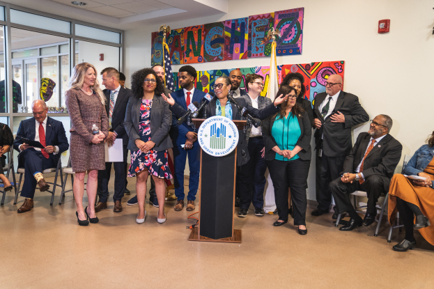 Eine Gruppe von Menschen steht und sitzt in einem Raum mit einem Podium, einem Mikrofon und einer Tafel mit der Aufschrift "Sancho-Stadtrat tritt dem Stadtrat bei", mit Flaggen, einem Feuerlöscher und Deckenleuchten an der Rückwand.
