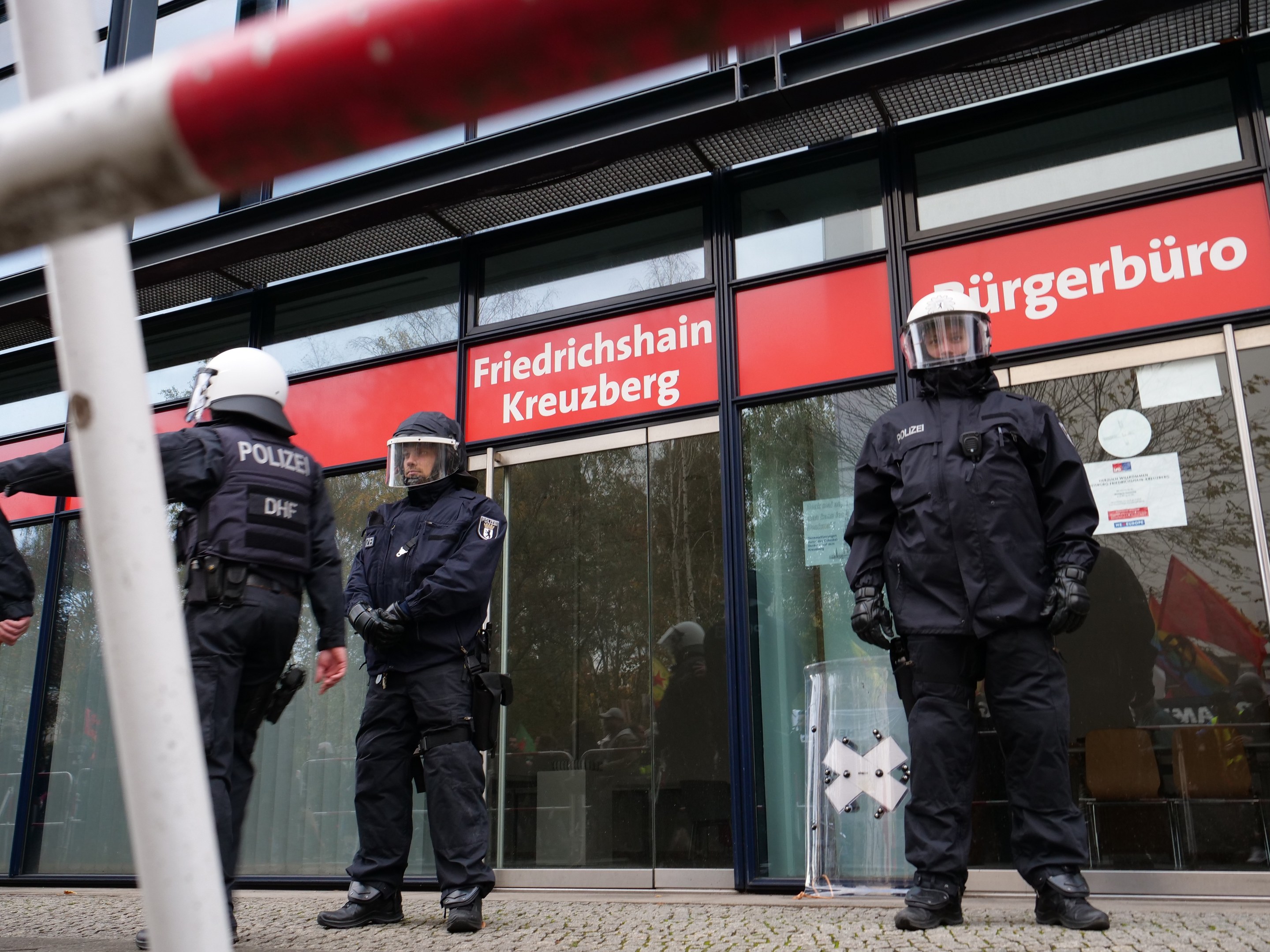 Polizeibeamte in Uniform stehen vor einem Glasfrontgebäude, tragen Helme und Handschuhe, mit einer Stange links und Schildern mit der Aufschrift "Friedrichshain Kreuzberg" im Hintergrund.