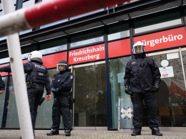 Polizeibeamte in Uniform stehen vor einem Glasfrontgebäude, tragen Helme und Handschuhe, mit einer Stange links und Schildern mit der Aufschrift "Friedrichshain Kreuzberg" im Hintergrund.