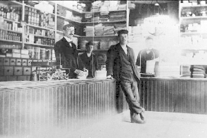 Drei Männer stehen an einem Tresen in einem Laden mit verschiedenen Gegenständen, ein buchgefülltes Regal im Hintergrund, während eines Raubüberfalls.