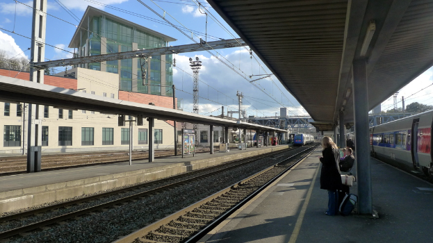Eine Bahnhofszenensz mit Personen, die rechts stehen und sitzen, einem fahrenden Zug in der Mitte und Gebäuden, Polen und einem bewölkten Himmel im Hintergrund.