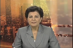 Eine Frau in einem Businessanzug sitzt vor einem Fernseher, im Hintergrund sind ein paar Gebäude zu sehen, die darauf hindeuten, dass sie eine Nachrichtensprecherin ist.