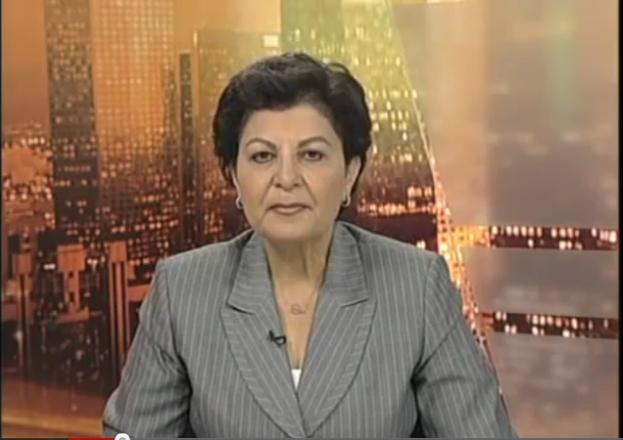 Eine Frau in einem Businessanzug sitzt vor einem Fernseher, im Hintergrund sind ein paar Gebäude zu sehen, die darauf hindeuten, dass sie eine Nachrichtensprecherin ist.