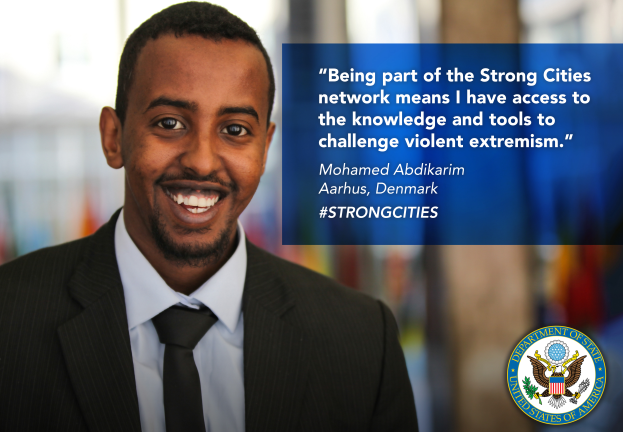 Ein Mann im Anzug und Krawatte, der strahlend in die Kamera lächelt, mit einem Logo und Text auf der rechten Seite, der besagt: "Teil des starken Städtenetzwerks zu sein bedeutet, Zugang zu Wissen und Werkzeugen zu haben, um gewalttätigen Extremismus in Frage zu stellen", und einem unscharfen Hintergrund.