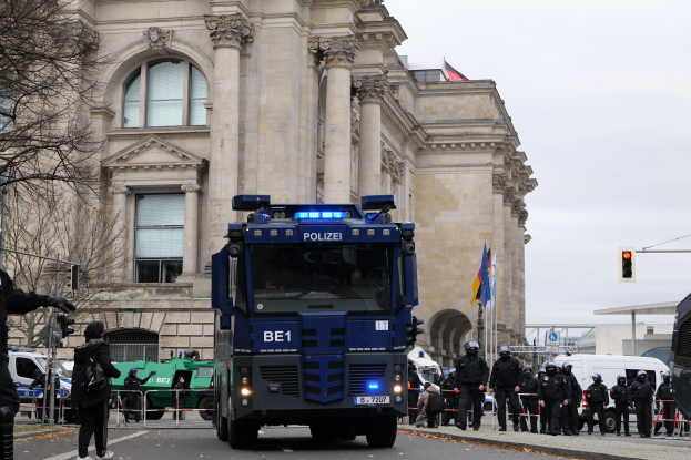 Eine Gruppe von Polizisten steht vor einem großen Gebäude mit Fenstern, Säulen und Bögen, mit Fahrzeugen auf der Straße, einer Person mit einer Kamera auf der linken Seite und Bäumen, Verkehrszeichen, Flaggen und einem klaren blauen Himmel im Hintergrund.