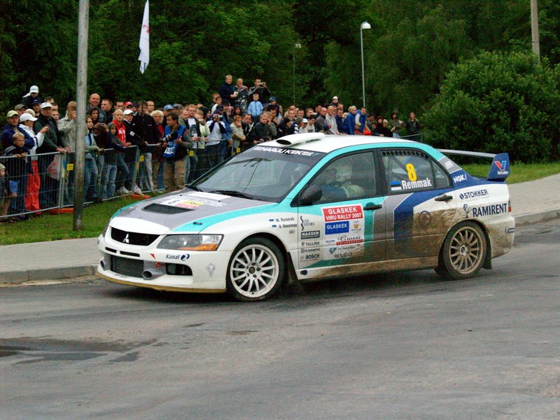 Ein Rallyeauto mit Text und Logos fährt an einer Menge vorbei, die Kameras hält, entlang einer Straße, die von Geländern, Fahnenmasten und Bäumen gesäumt ist.