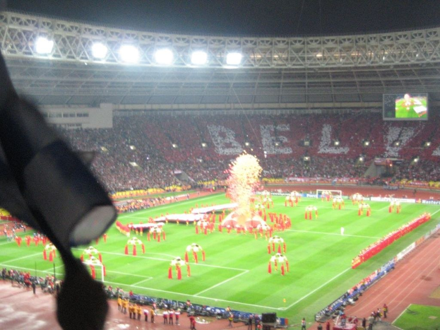 Ein Stadion mit Flutlicht, Eisengittern, Zuschauern, Menschen am Boden, Werbetafeln, Anzeigetafeln, Überwachungskamera, Fahnen und Dekorationen.