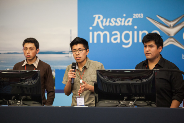 Drei Jungen stehen neben einem Schreibtisch mit zwei Computersystemen, einer spricht in ein Mikrofon, tragen ID-Karten mit roten Tags, vor einer Wand mit einer Werbetafel, auf der "Russia Imagine 2013" steht.