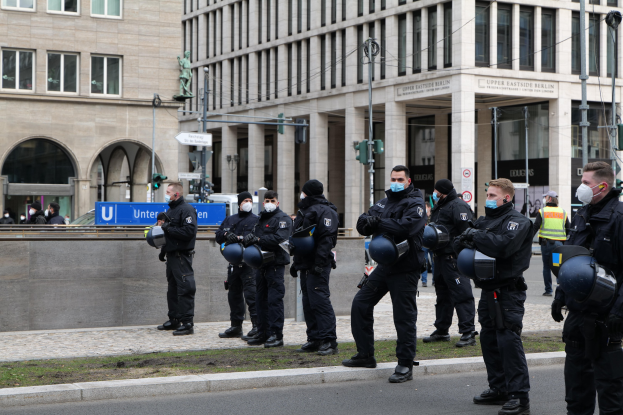 Polizeibeamte in schwarzen Uniformen und Masken stehen vor einem Berliner Gebäude mit Glasfenstern und Säulen, einige halten Helme, mit Laternen, Verkehrsampeln, Schildern mit Text, einer Statue und anderen Menschen in der Szene.